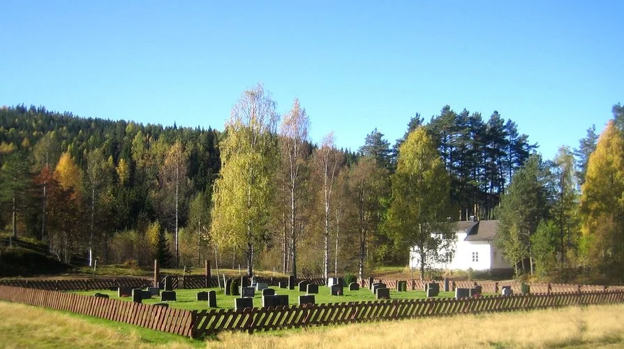 Luksefjell kirkegård med gravminner og gjerdet rundt. Hus i bakgrunnen. Foto.